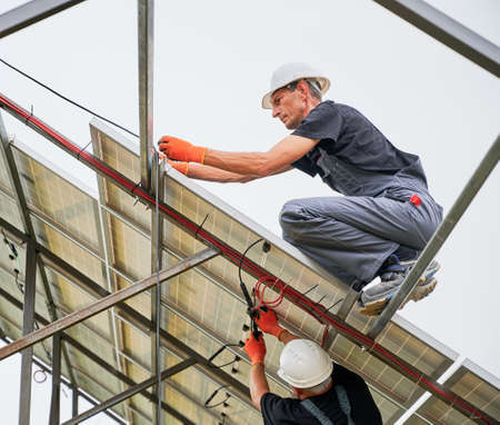Two men in safety construction helmets installing photovoltaic solar panel system under white sky. Male workers building solar modules, panels and support structures of photovoltaic solar array.の写真素材
