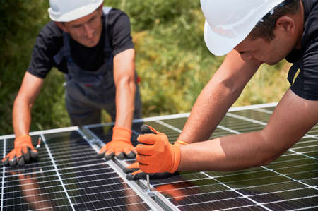 Male workers building photovoltaic solar panel system outdoors. Men engineers placing solar module on metal rails, wearing construction helmets and work gloves. Renewable and ecological energy.の写真素材