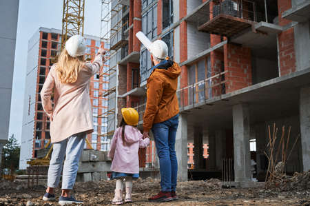 Back view of man and woman in safety helmets holding daughter hands and pointing at tower crane at construction site. Family with kid standing outside building under construction.の写真素材