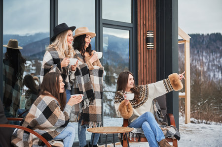 Young women enjoying winter weekends on terrace of contemporary barn house. Four girls having fun, sitting on chairs and drinking hot tea.の写真素材