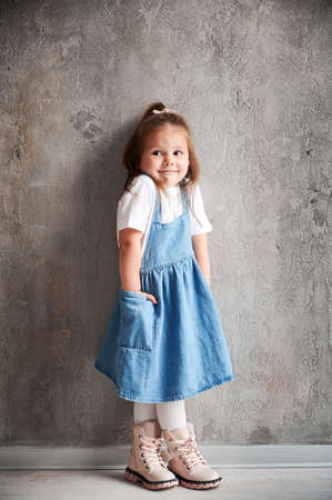 Full length of adorable little girl in denim dress standing against the wall in new home. Cute female child keeping hand in pocket.の写真素材
