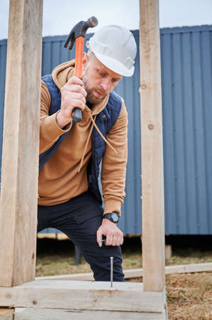 Man worker building wooden frame house on pile foundation. Carpenter hammering nail into wooden...