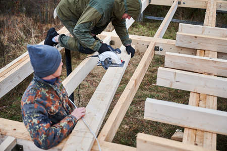 Carpenter using circular saw for cutting wood beam. Man worker building wooden frame house on pile...