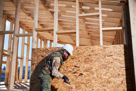 Carpenter hammering nail into OSB panel on the wall of future cottage. Man worker building wooden frame house. Carpentry and construction concept.の写真素材