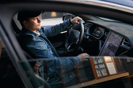 View through open side car window. Man in cap sitting in elite automobile, putting one hand on steering wheel and by other hand entering destination address on large touch screen.の写真素材