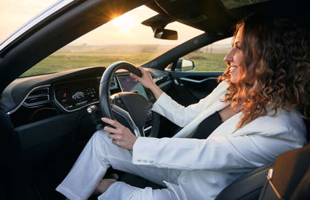 Beautiful young woman in elegant white suit holding steering wheel and smiling while sitting in...