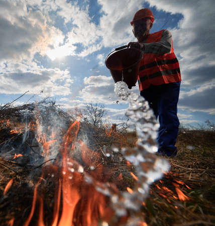 Fireman ecologist extinguishing fire in field with cloudy sky on background. Low angle view male environmentalist holding bucket and pouring water on burning dry grass. Natural disaster concept.の写真素材