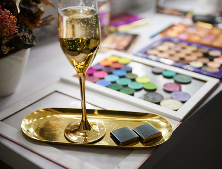 Dressing table with makeup eyeshadow palette, glass of alcoholic drink and corporate business chocolate on gold tray. Champagne, sweets and makeup tools in visage studio.の写真素材