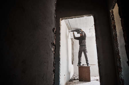 Full length of man builder standing on wooden table and using electric drill while making hole in...
