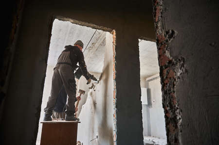 Full length of male builders using hammer drill and sledgehammer while destroying wall in building....