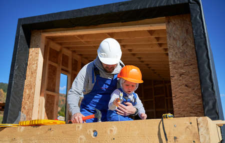 Father with toddler son building wooden frame house. Male builders hammering nail into plank on construction site, wearing helmet and blue overalls on sunny day. Carpentry and family concept.の写真素材