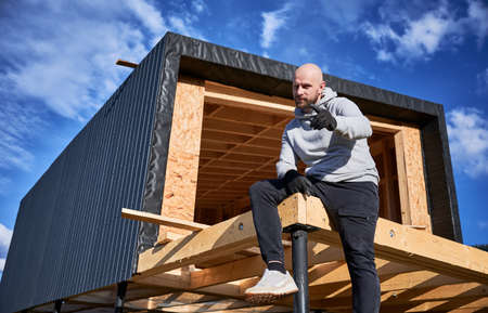 Male developer building wooden frame house on pile foundation. Portrait of bald man sitting on terrace on construction site, inspecting quality of work.の写真素材