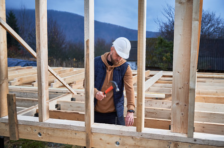Man worker building wooden frame house on pile foundation. Carpenter hammering nail into wooden joist, using hammer. Carpentry concept.の写真素材