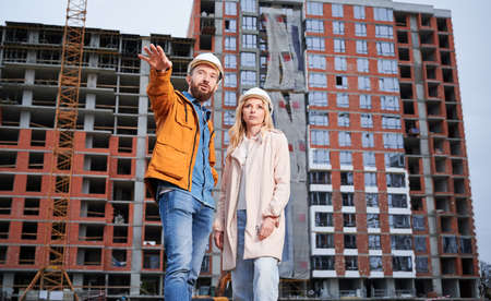 Couple in safety helmets standing outside apartment building under construction. Man and woman...