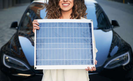 Cropped view of smiling woman holding solar panel on background of black electric car. Concept of environmental energy for automobiles powered by battery-powered electric motors.の写真素材