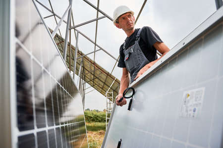 Serious man solar installer assembling photovoltaic solar panel system outdoors. Male worker wearing safety construction helmet and work overalls while standing by solar modules.の写真素材