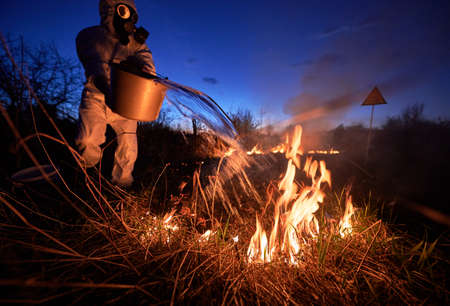 Research scientist extinguishing fire in field with blue night sky on background. Man in protective radiation suit and gas mask holding bucket and pouring water on burning grass with smoke.の写真素材