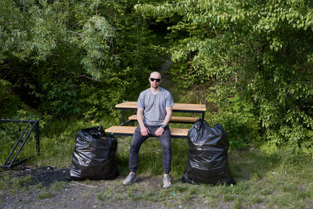 Young man in sunglasses sitting on wooden bench near garbage bags. Male person resting in forest...
