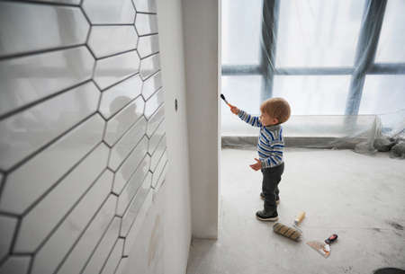Side view of adorable child painting wall in room under renovation. Cute kid in striped sweater...