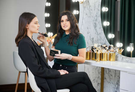 Portrait of beauty specialist applying cosmetics on woman face in visage studio. Stylish woman...