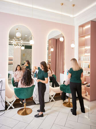 Two female hairstylists curling client hair and doing hairstyle in modern beauty salon. Young women sitting in chairs in front of mirrors while hairdressers styling clients hair.の写真素材