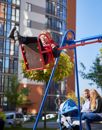 Little son having fun on swing, while mum and dad sitting on bench with pram at playground. Side...