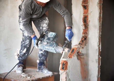 Close up of male builder in workwear drilling wall with hammer drill. Man worker using drill breaker while destroying wall in apartment under renovation. Demolition work and home renovation concept.の写真素材