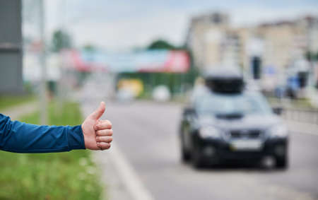 Close up of mans hand hitchhiking by roadside. Male hand showing thumbs up gesture outdoors on blurred background. Hitchhiking, hitching, auto stop concept.の写真素材