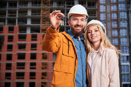 Happy couple with new home keys standing on the street with apartment building under construction on background. Smiling man homeowner hugging woman at construction site.の写真素材
