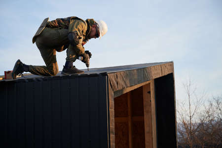 Male builder installing black corrugated iron sheet used as facade of future cottage. Man worker building wooden frame house. Carpentry and construction concept.の写真素材