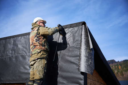 Male builder installing waterproof membrane on the wall of future cottage. Man worker building wooden frame house with blue sky on background. Carpentry and construction concept.の写真素材