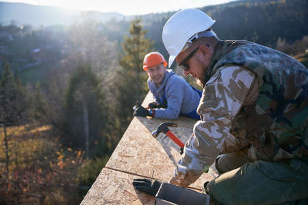 Carpenter hammering nail into OSB panel on the roof top of future cottage. Man worker building wooden frame house. Carpentry and construction concept.の写真素材