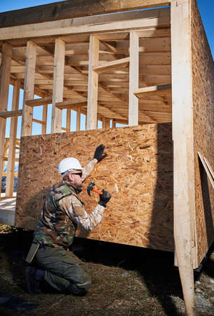 Carpenter hammering nail into OSB panel on the wall of future cottage. Man worker building wooden...