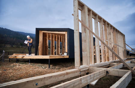 Male architect building wooden frame house in the Scandinavian style barnhouse. Man builder standing on construction site in safety helmet, with construction documentation inspecting quality of work.の写真素材