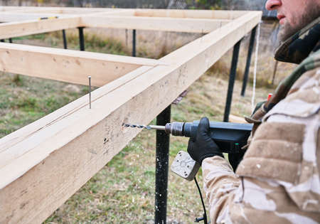 Man building wooden house on pile foundation. Close up of male worker drilling hole by electric drill in wooden frame of future house on construction site. Carpentry concept.の写真素材