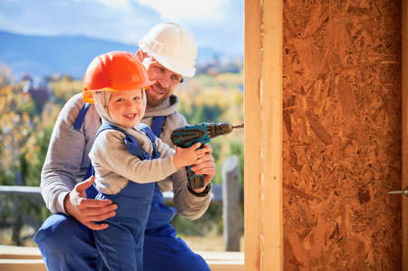 Father with toddler son building wooden frame house. Male builder and kid using screwdriver on...