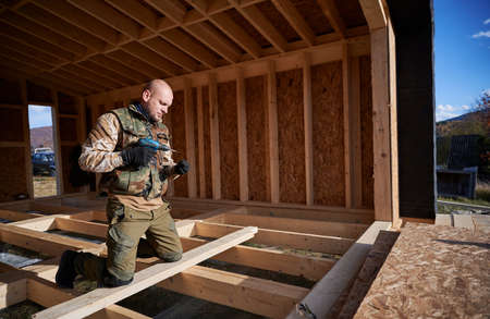 Male builder working with screwdriver inside of future cottage. Man worker building wooden frame...
