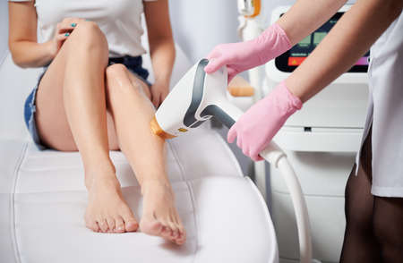 Close up of beautician in sterile gloves removing hair on female leg with laser epilation machine. Cosmetologist using diode hair removal device while performing epilation procedure on woman leg.の写真素材