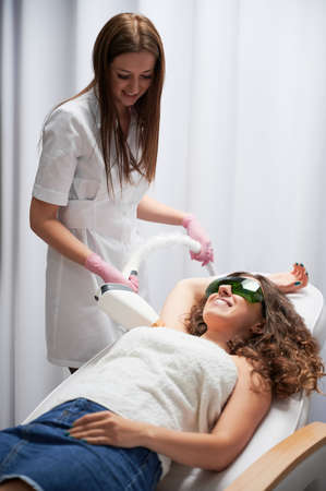 Laser skin care. Cosmetologist doing hair removal from axillary area of happy client. Young woman with smile expression liying on white couch and enjoying pleasant procedure of losing unwanted hair.の写真素材