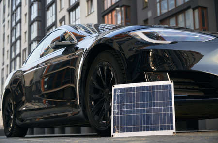 Black electric vehicle and photovoltaic solar panel on city street. Portable solar panel and modern...