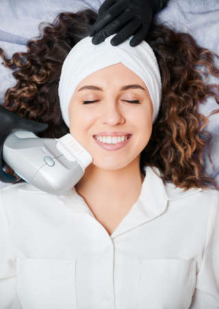 Top view of beautiful young woman with closed eyes wearing white headband while receiving facial treatment at wellness center. Cheerful female patient having face lifting procedure and smiling.の写真素材
