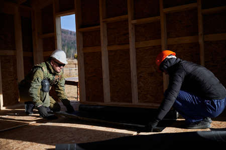 Male builders installing waterproof membrane on the wall of future cottage. Men workers building...