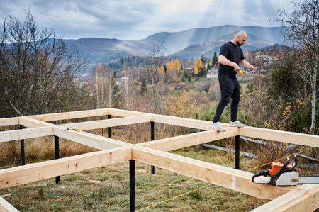 Man worker building wooden frame house on pile foundation in the mountains. Carpenter measuring...