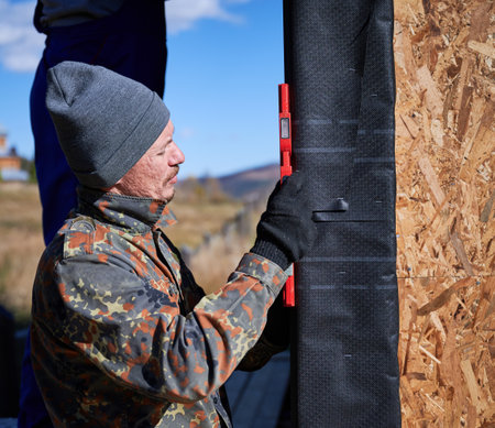 Male builders using red spirit level and measuring vertical surface of new wall, installing...