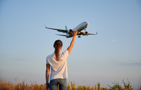 Back view of woman waving hand to flying commercial airplane in the sky at sunset. Lifestyle and travel concept.の写真素材