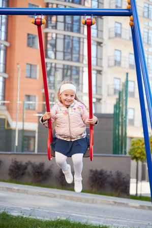 Little female child, wearing warm clothes, swaying on swing, having fun at playground. Front view of adorable girl on swing smiling, looking at camera, while swinging outdoors. Concept of joy.の写真素材
