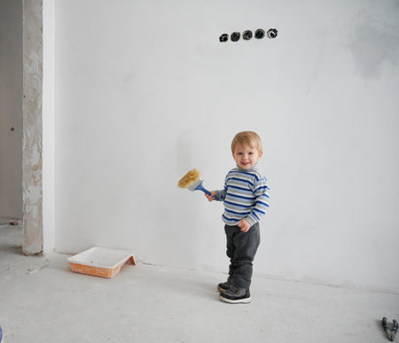 Adorable little boy looking at camera and smiling while holding paint brush. Joyful male kid with paint tool in his hand standing against white wall in apartment under renovation.の写真素材