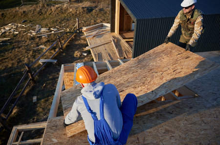 Carpenters mounting wooden OSB board on rooftop of future cottage. Men workers building wooden frame...