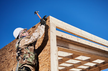 Carpenter hammering nail into OSB panel on the wall of future cottage. Man worker building wooden frame house. Carpentry and construction concept.の写真素材