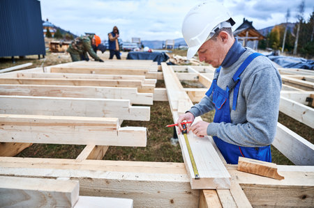Man worker building wooden frame house on pile foundation. Carpenter using tape measure for...